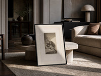 Man with grotesque mask figures print in a black frame leaning on a travertine table in a sunlit lounge