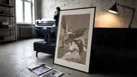 Winged demon carrying frightened figure print in a slim black frame leaning on a dark sofa in a concrete print studio