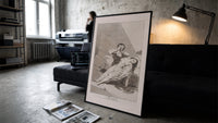 Man grieving over collapsed woman print in a slim black frame leaning on a dark sofa in a concrete print studio