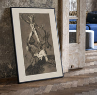 Grotesque figures climbing atop one another print in a black frame leaning beside a vintage door on parquet