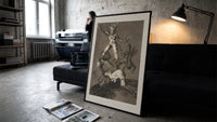 Grotesque figures climbing atop one another print in a slim black frame leaning on a dark sofa in a concrete print studio