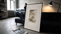 Grotesque figures carrying woman aloft print in a slim black frame leaning on a dark sofa in a concrete print studio