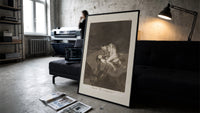 struggling figures with flying witches print in a slim black frame leaning on a dark sofa in a concrete print studio