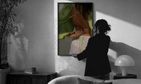 Green Tie-Back Blouse and White Trousers print in a slim black frame being hung on a sunlit wall in a monochrome room