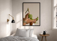 Green Woven Stiletto Sandals print in a black frame above a bed with striped linens, a brass wall lamp and a sunlit window