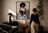 Hair Braiding Scene with Two Figures print in a black frame on a sunlit wall with a viewer
