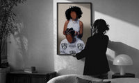 Hair Braiding Scene with Two Figures print in a slim black frame being hung on a sunlit wall in a monochrome room
