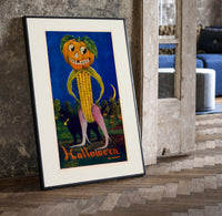 Anthropomorphic pumpkin-headed corn figure print in a black frame leaning beside a vintage door on parquet