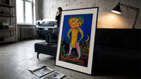 Anthropomorphic pumpkin-headed corn figure print in a slim black frame leaning on a dark sofa in a concrete print studio