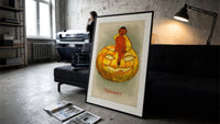 Red devil sitting on jack-o-lantern print in a slim black frame leaning on a dark sofa in a concrete print studio