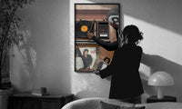 Hand on a Wooden Record Player print in a slim black frame being hung on a sunlit wall in a monochrome room