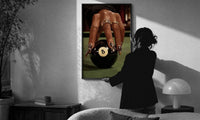 Hand on the Eight Ball print in a slim black frame being hung on a sunlit wall in a monochrome room