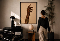Hand with a Cross and Gold Chains print in a black frame on a sunlit plaster wall, a woman looking on beside a mushroom lamp