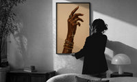 Hand with a Cross and Gold Chains print in a slim black frame being hung on a sunlit wall in a monochrome room