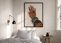Hand with Blue Bangles print in a black frame above a bed with striped linens, a brass wall lamp and a sunlit window