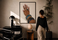 Hand with Blue Bangles print in a black frame on a sunlit plaster wall, a woman looking on beside a mushroom lamp