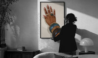 Hand with Blue Bangles print in a slim black frame being hung on a sunlit wall in a monochrome room
