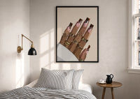 Hand with Brown Swirl Nails print in a black frame above a bed with striped linens, a brass wall lamp and a sunlit window