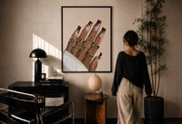 Hand with Brown Swirl Nails print in a black frame on a sunlit plaster wall, a woman looking on beside a mushroom lamp