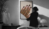 Hand with Brown Swirl Nails print in a slim black frame being hung on a sunlit wall in a monochrome room