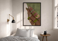 Hand with Green Nails and Rings print in a black frame above a bed with striped linens, a brass wall lamp and a sunlit window