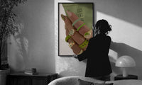 Hand with Green Nails and Rings print in a slim black frame being hung on a sunlit wall in a monochrome room