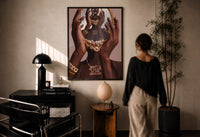 Hands Holding an Ornate Mirror print in a black frame on a sunlit plaster wall, a woman looking on beside a mushroom lamp