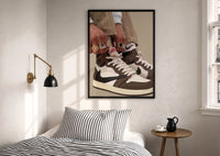 Hands Lacing Brown High-Tops print in a black frame above a bed with striped linens, a brass wall lamp and a sunlit window