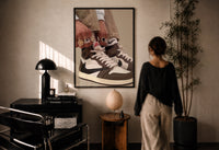 Hands Lacing Brown High-Tops print in a black frame on a sunlit plaster wall, a woman looking on beside a mushroom lamp