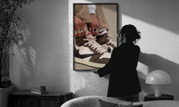 Hands Lacing Brown High-Tops print in a slim black frame being hung on a sunlit wall in a monochrome room