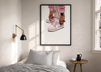 Pink Low-Tops print in a black frame above a bed with striped linens, a brass wall lamp and a sunlit window