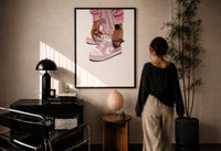 Pink Low-Tops print in a black frame on a sunlit plaster wall, a woman looking on beside a mushroom lamp
