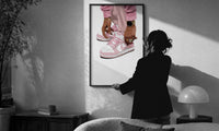 Pink Low-Tops print in a slim black frame being hung on a sunlit wall in a monochrome room