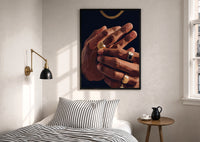 Hands with Gold Rings on Navy print in a black frame above a bed with striped linens, a brass wall lamp and a sunlit window