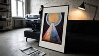 Golden sun with pyramid of colors print in a slim black frame leaning on a dark sofa in a concrete print studio