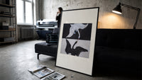 Two swans, one white one black print in a slim black frame leaning on a dark sofa in a concrete print studio
