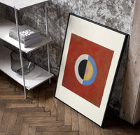 Divided circle with concentric rings poster in a black frame leaning beside a shelf in a raw-plaster loft