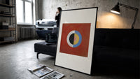Divided circle with concentric rings print in a slim black frame leaning on a dark sofa in a concrete print studio