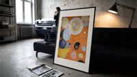 Abstract spirals and snail shells print in a slim black frame leaning on a dark sofa in a concrete print studio