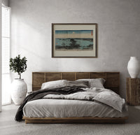 Japanese bay landscape with moon print in a dark wood frame above a rattan bed on a plaster wall