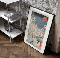 Snowy bridge with travelers in winter poster in a black frame leaning beside a shelf in a raw-plaster loft
