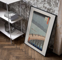 People crossing bridge in rain poster in a black frame leaning beside a shelf in a raw-plaster loft