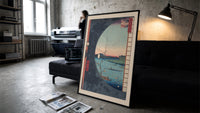 River landscape with plum blossom branch print in a slim black frame leaning on a dark sofa in a concrete print studio