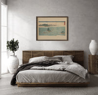 Japanese whirlpools and coastal seascape print in a dark wood frame above a rattan bed on a plaster wall
