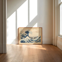 Great wave with boats and Mount Fuji print in an oak frame on a sunlit wood floor beneath tall window light