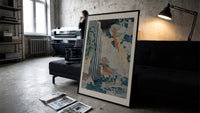 Japanese waterfall with travelers and hut print in a slim black frame leaning on a dark sofa in a concrete print studio