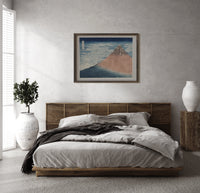 Mount Fuji with snow-capped peak print in a dark wood frame above a rattan bed on a plaster wall