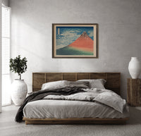 Red Mount Fuji with clouds print in a dark wood frame above a rattan bed on a plaster wall