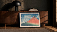 Red Mount Fuji with clouds print in a black frame leaning against a walnut console in lamplight