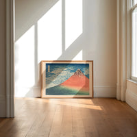 Red Mount Fuji with clouds print in an oak frame on a sunlit wood floor beneath tall window light